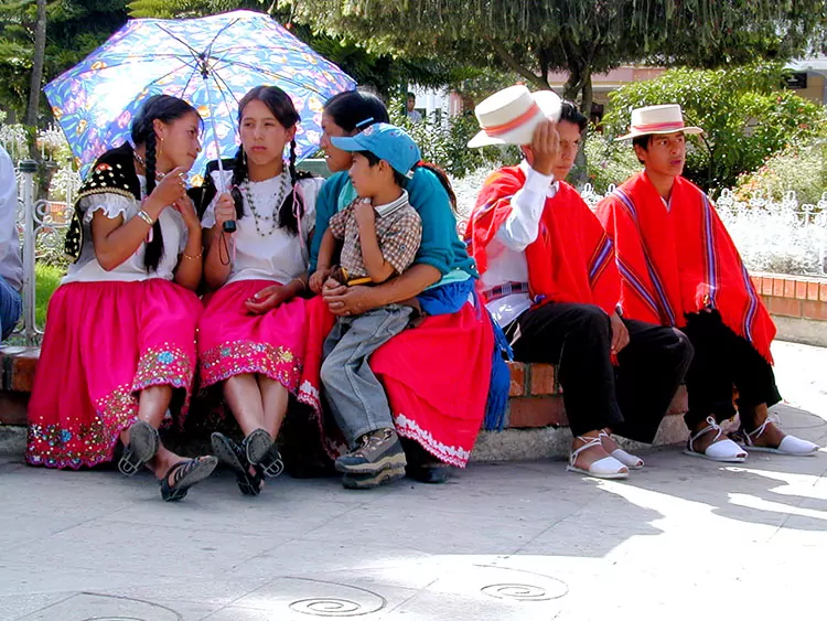 Women and men dressed in colorful local attire sit in a park in Cuenca