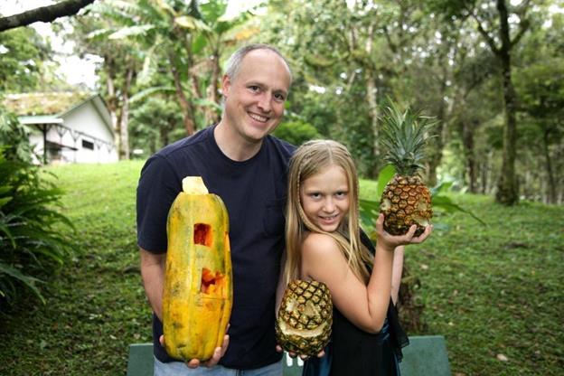 Father and daughter pose with tropical halloween carvings. One is a halloween pineapple. Palm trees are in the background.