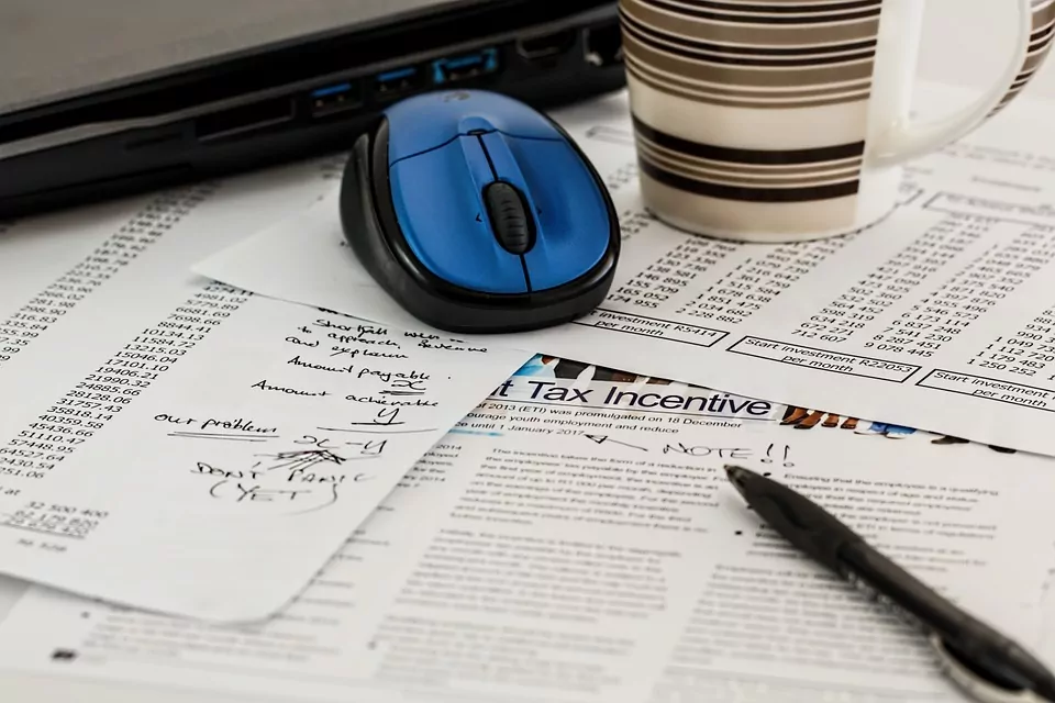 pile of tax papers with a pen, mouse, coffee mug and laptop on top of them