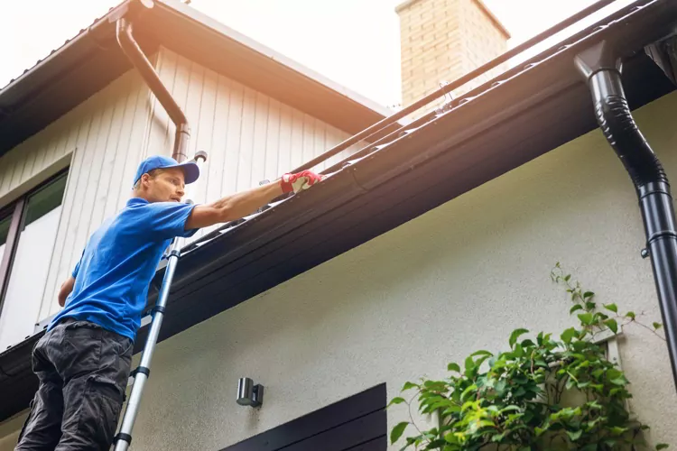 Man on ladder cleaning house gutter from leaves and dirt