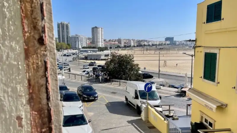 Cars parked along a narrow street with a sandy beach, tall buildings, and a clear blue sky in the background.