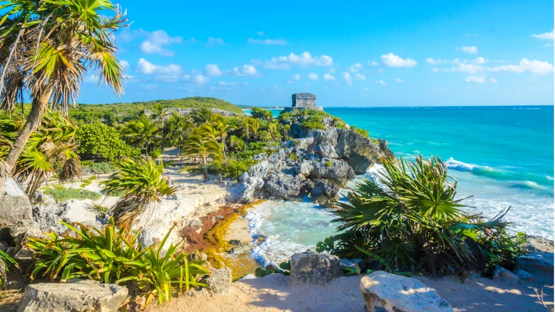 A panoramic view of the ancient Mayan ruins of Tulum perched on a cliff overlooking the turquoise Caribbean Sea, showcasing the natural beauty that drives Real Estate in Tulum.