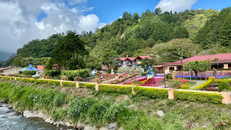 The landscape is lush and green, with forest-covered hills in the background. In the middle, there’s a well-maintained park featuring colorful flower beds arranged in patterns, decorative structures, and small buildings with red roofs.