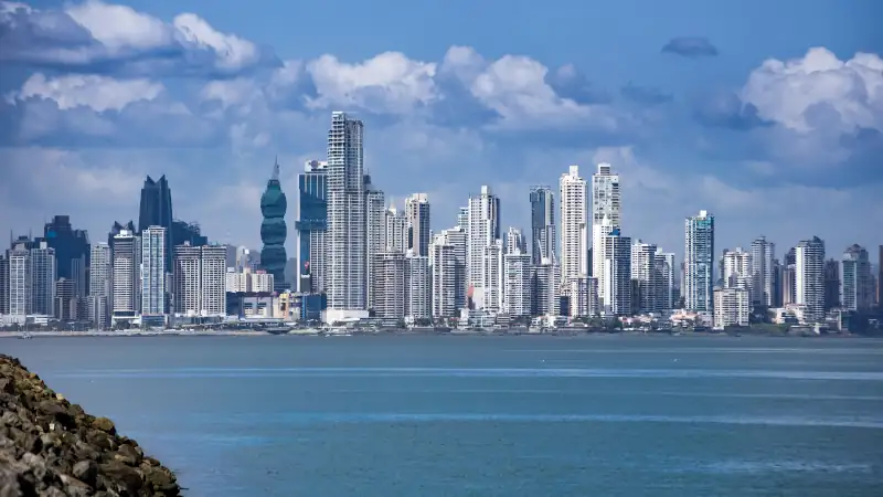 Skyline of Panama City with modern skyscrapers along the ocean under a blue sky with clouds.