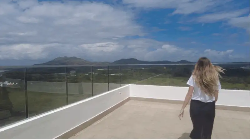 Sophia walks across a modern rooftop terrace overlooking a scenic mountain landscape under a bright blue sky with scattered clouds. The open setting and panoramic view create a calm, airy atmosphere.