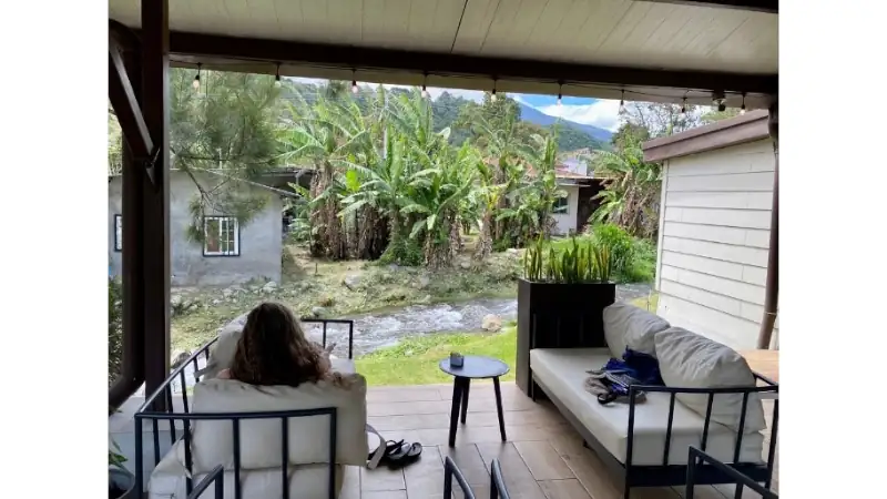 A cozy covered patio with cushioned seating overlooks a small stream, lush banana trees, and misty mountain scenery, creating a peaceful tropical retreat.