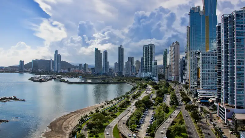 Panoramic view of Panama City's modern skyline along the waterfront, with high-rise buildings, a coastal boulevard, and dramatic clouds overhead.