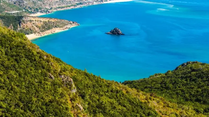 Panoramic view of a turquoise bay surrounded by lush green hills and rugged cliffs, with a small rocky islet rising from the crystal-clear water.
