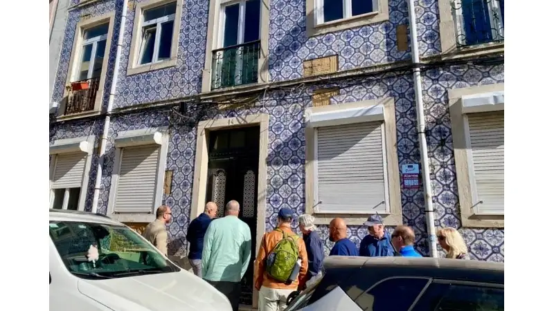 A group of people standing outside a historic blue-tiled residential building, gathered near the entrance on a sunny street.