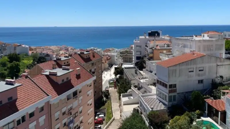 A panoramic view of a coastal neighborhood with red-roofed buildings overlooking the ocean under a clear blue sky.