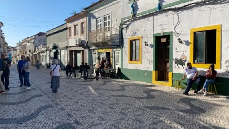 Historic pedestrian street lined with colorful buildings, cafés, and traditional black-and-white mosaic paving under a clear blue sky.