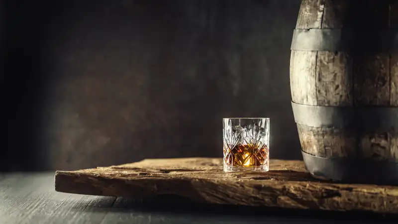 Glass of whisky or bourbon in ornamental glass next to a vinatge wooden barrel on a rustic wood and dark background