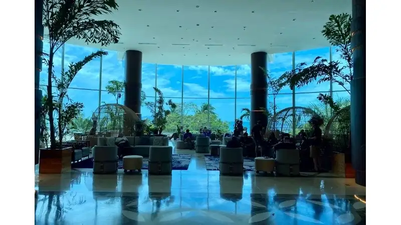 A spacious modern hotel lobby featuring floor-to-ceiling windows, elegant lounge seating, tropical plants, and panoramic views of lush greenery beneath a bright blue sky.