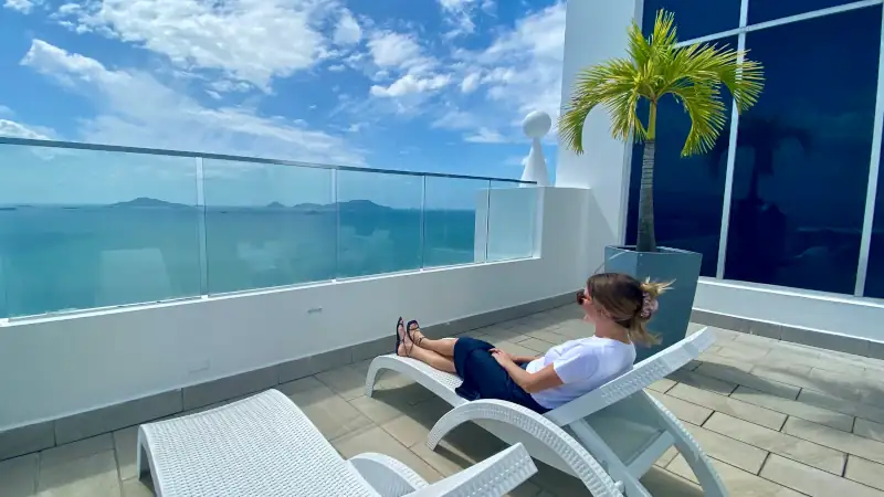 Sophia relaxes on a sun lounger on a modern rooftop terrace overlooking the turquoise ocean and distant islands, beneath a bright blue sky.